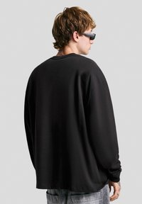 Un jeune homme portant des lunettes de soleil, vêtu d'une chemise noire oversized à manches longues et d'un jean gris clair, tourné légèrement sur le côté devant un fond uni.