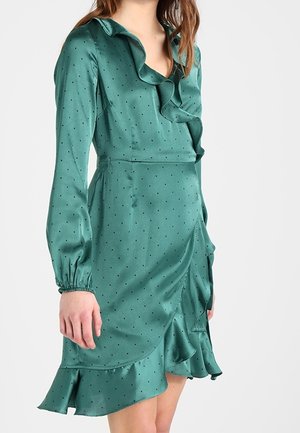 Kobieta w turkusowej sukience kopertowej z satyny z czarnymi groszkami, długimi rękawami, falbankowym kołnierzem i falbaną na dole.