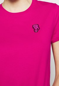 Camiseta de color rosa brillante con un pequeño pin de personaje de dibujos animados en tono rosa oscuro en el lado izquierdo del pecho, que muestra a una persona con gafas de sol y coleta.