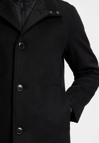 Cappotto in lana nero con bottoni, colletto a punta, bottoni dorati sulla patta del colletto e una mano inserita nella tasca laterale di una persona.