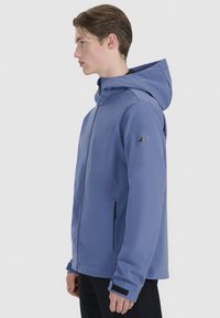 Jeune homme debout de profil, portant une veste à capuche bleu clair avec fermeture éclair devant et un pantalon noir, sur un fond uni clair.