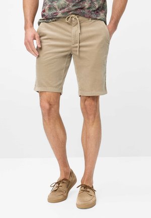 Man draagt beige corduroy shorts met trekkoord in de taille en lichtbruine casual veterschoenen, staand met handen gedeeltelijk in de zakken.