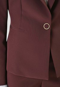 Blazer bordeaux in tessuto intrecciato, con bottone circolare color oro, rever a lancia e struttura elegante dal design minimalista.