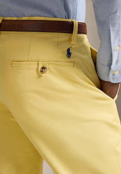 Κίτρινο παντελόνι από βαμβάκι με λεία υφή. Διαθέτει πίσω τσέπη με κουμπί και μπλε λογότυπο. Ορατή καφέ ζώνη.