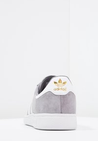 Szary sportowy but zamszowy z trzema białymi paskami, białą gumową podeszwą i złotym logo Adidas na zapiętku. Widok z tyłu.