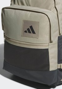 Mochila de tela beige con una sección inferior de malla negra, que presenta un parche de logo rectangular con tres franjas negras y compartimentos con cremallera.