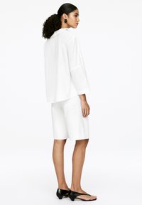 White, long-sleeve shirt with a ruffled collar, paired with white shorts. Black, flat sandals. Smooth fabric, minimalistic design.