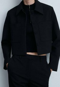 Veste noire raccourcie avec col pointu, deux poches avant et fermeture à boutons ; associée à un haut noir côtelé et un pantalon assorti.