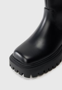 Mustam nahast pahkluu saapad ümara varbaga, paksu struktuurse kummitaldaga ja väljendunud mustriga põhjal, mis tagab haarde.