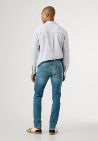 Camisa de botones a rayas azul claro, jeans ajustados azul oscuro, zapatillas blancas con acentos en azul marino y rojo, vistas desde la parte de atrás.