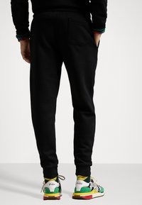 Černé tepláky s úzkým střihem, s kapsou na zadní straně. Kombinovány s vícebarevnými teniskami a vzorovanými ponožkami.