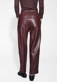 Pantaloni in pelle bordeaux con una texture liscia, vita elastica e due tasche posteriori, caratterizzati da un design a gamba dritta e cuciture minimal.