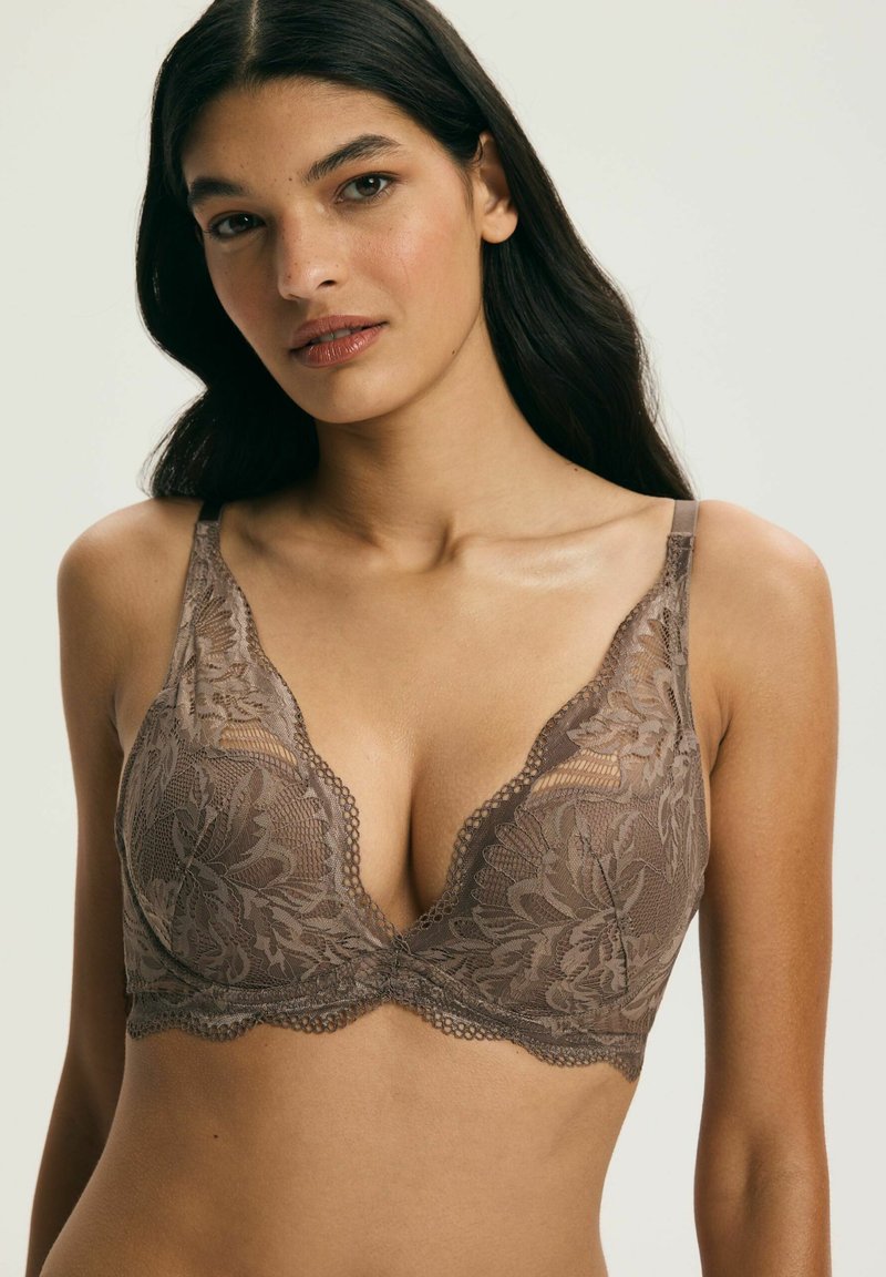 Kobieta ubrana w brązowy koronkowy biustonosz z kwiatowymi wzorami, cienkimi ramiączkami i falistymi krawędziami na tle jasnego, jednolitego tła.