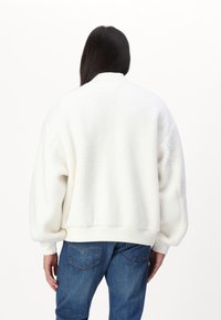 Persoon met lang donker haar, gekleed in een witte fleece jas en blauwe spijkerbroek, met de rug naar de camera gekeerd tegen een effen witte achtergrond.