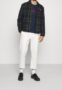 Man klädd i vita korta byxor, svart och blå rutigt jacka, marinblå grafisk t-shirt, vita strumpor och svarta skor som står mot vit bakgrund.