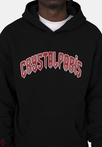 Sudadera negra con un gráfico de texto rojo "CRYSTALPARIS" en una fuente atrevida y arqueada. Incluye un bolsillo frontal y capucha con cordón.