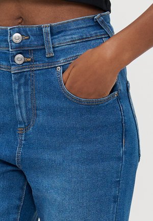 Main dans la poche avant d'un jean en denim bleu taille haute avec double boutons argentés et coutures visibles.