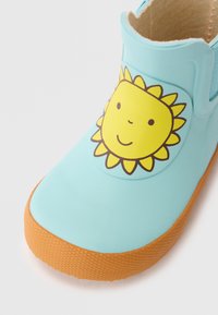 Botte en caoutchouc bleu clair avec un graphique de soleil. Elle présente une semelle orange texturée et des panneaux latéraux élastiques pour un enfilage facile. Fini lisse dans l'ensemble.