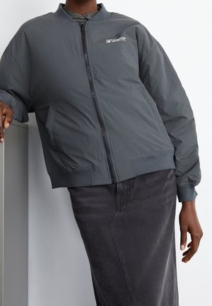 Giacca bomber grigia con zip frontale, polsi e orlo a costine. Presenta un logo bianco sul petto e un tessuto testurizzato. Abbinata a una gonna scura.