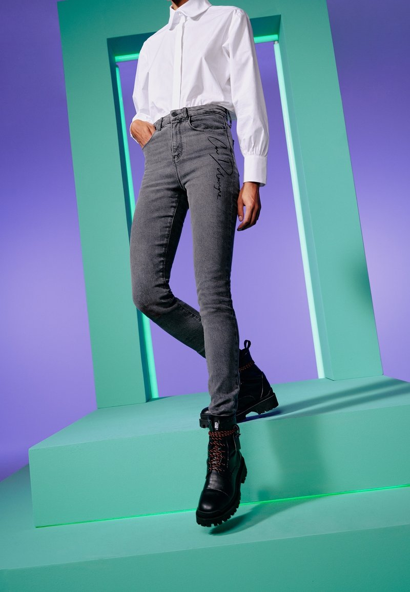 Jean skinny gris avec broderie sur le côté, porté avec une chemise blanche à col et des bottes noires à lacets, présenté devant un fond coloré.