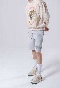 Persona lleva un sudadera beige de Ed Hardy con un gráfico de tigre, pantalones cortos grises con cordón, calcetas blancas y zapatillas beige con suelas verdes.