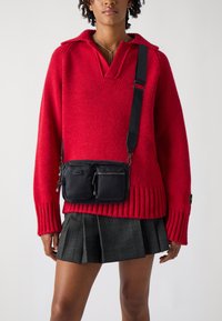 Pull en tricot rouge avec un col en V, des poignets côtelés, associé à un sac en nylon noir en bandoulière avec deux poches zippées. Mini-jupe à carreaux gris.