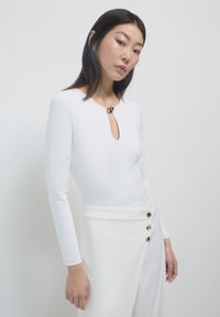 Bodysuit blanc à manches longues avec un décolleté en forme de trou de serrure, doté d'un accent métallique rond au cou, associé à un pantalon blanc taille haute.
