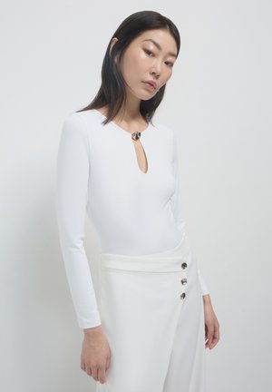 Bodysuit blanc à manches longues avec un décolleté en forme de trou de serrure, doté d'un accent métallique rond au cou, associé à un pantalon blanc taille haute.