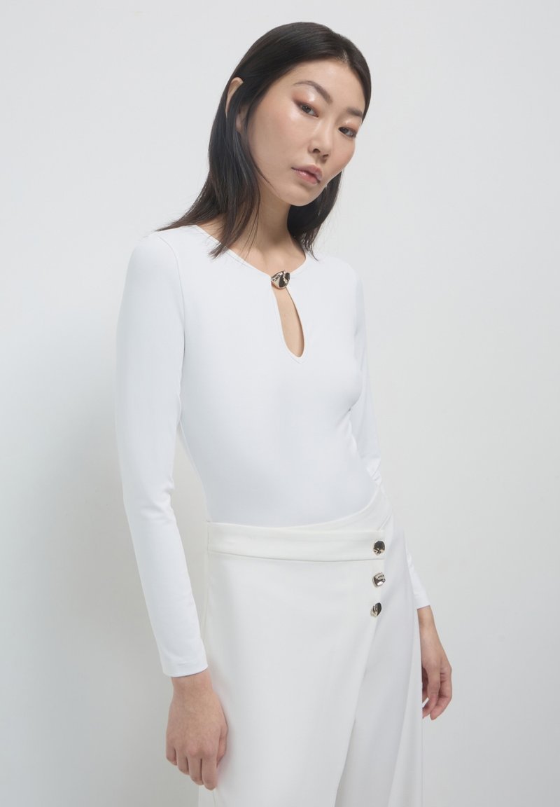 Bodysuit blanc à manches longues avec un décolleté en forme de trou de serrure, doté d'un accent métallique rond au cou, associé à un pantalon blanc taille haute.