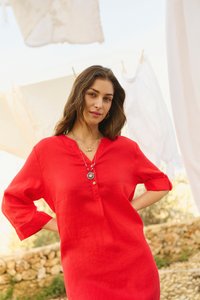Femme en robe rouge aux manches retroussées et aux colliers superposés debout à l'extérieur près de linges blancs suspendus par une journée ensoleillée.