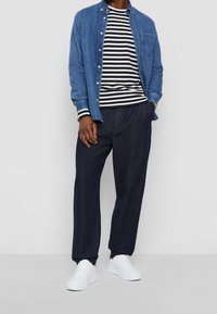 Jeansskjorta med knappar över en svart- och vitrandig långärmad skjorta, tillsammans med mörkblå vida byxor och vita sneakers.