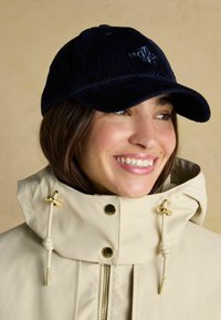 Jūras spēku krāsas korduroja cepure ar izliekto tempu un izšūtu logotipu, valkāta ar ziloņkaula krāsas jaku, kurai ir zelta toņa aksesuāri un aukla.