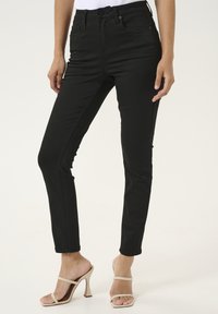 Schwarze, hochgeschnittene Skinny Jeans aus dehnbarem Stoff. Verfügt über fünf Taschen, Gürtelschlaufen und eine glatte Oberfläche. Kombiniert mit beigen Absatzsandalen.