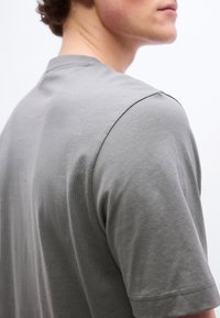 Νεαρό άτομο που φοράει απλό γκρι κοντομάνικο μπλουζάκι, εμφανίζεται από πίσω και πλάγια, με κατσαρά μαλλιά και ουδέτερο φόντο.