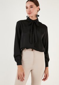 Bluză neagră, cu mâneci lungi și guler legat, realizată dintr-un material neted. Purtați-o cu pantaloni bej, ajustați, evidențiind un design simplu și elegant.