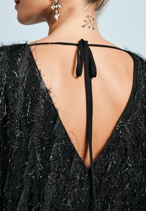 Abito nero con scollatura a V profonda sul retro, realizzato in tessuto testurizzato con fili metallici e chiusura a fiocco sottile nera. Orecchini di cristallo visibili.