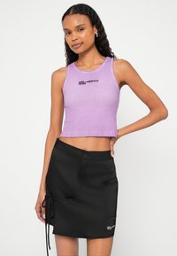 Žena v purpurovom bezrukávovom crop tope a čiernej mini sukni, pózuje s jednou rukou na bedre na jednoduchom bielom pozadí.