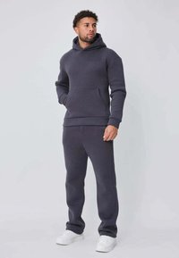 Sweat à capuche gris foncé et jogging assorti en matériau doux, avec une poche avant, une texture brossée et des poignets côtelés, assortis à des baskets blanches.
