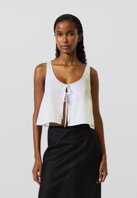 Ženska s kodrastimi lasmi, oblečena v brezrokavnik bel krojeni top s trakovi spredaj in visokim pasom črno krilo, na enostavni ozadju.