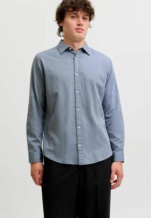Jeune homme aux cheveux bouclés portant une chemise bleu clair à boutons à manches longues et un pantalon noir, debout devant un fond blanc uni.