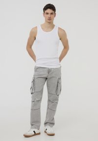 Top senza maniche bianco, pantaloni cargo grigi con tasche laterali e scarpe da ginnastica bianche con accenti neri. Modella in piedi contro uno sfondo neutro.