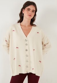 Cardigan crema con scollo a V e ricamo a forma di fiocco rosso, caratterizzato da grandi bottoni decorativi, vestibilità morbida e tessuto a maglia testurizzato.