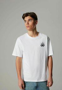 Vit bomull t-shirt med rund halsringning, korta ärmar och en svart grafisk design med två cirklar på bröstet. Ljusblå denimjeans.