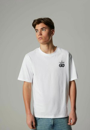 Vit bomull t-shirt med rund halsringning, korta ärmar och en svart grafisk design med två cirklar på bröstet. Ljusblå denimjeans.