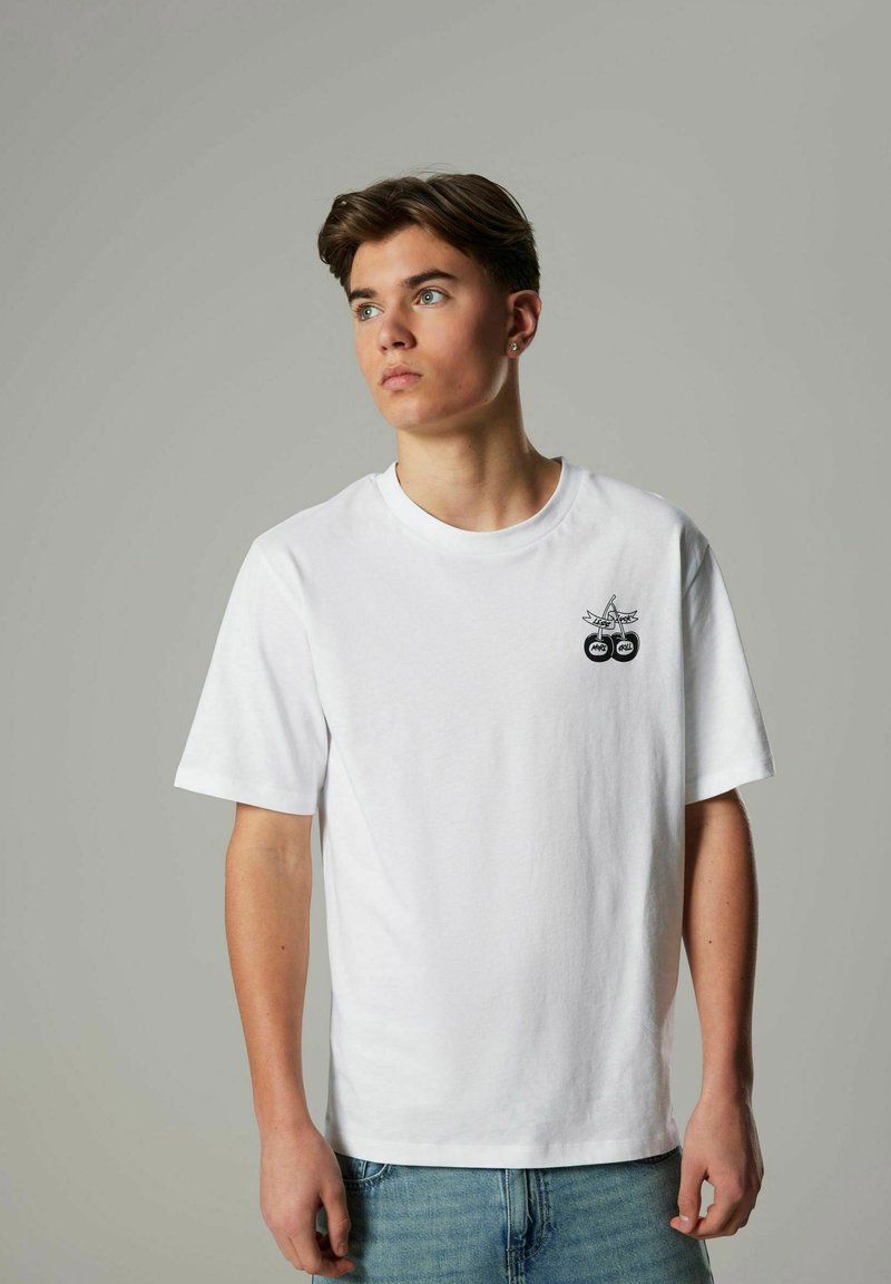 Vit bomull t-shirt med rund halsringning, korta ärmar och en svart grafisk design med två cirklar på bröstet. Ljusblå denimjeans.