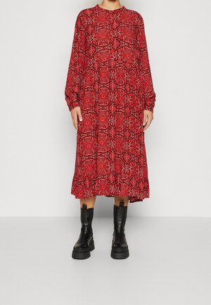 Person, die ein langärmeliges, rot geblümtes Midikleid trägt, kombiniert mit schwarzen klobigen Plateaustiefeln, steht vor einem schlichten Hintergrund.