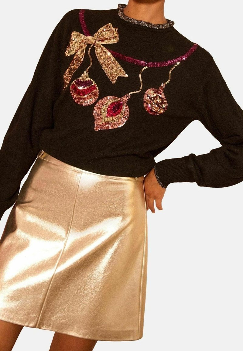Pull noir avec ornements de fête en sequins et nœud, associé à une jupe dorée brillante de style trapèze, porté par une personne la main sur la hanche.