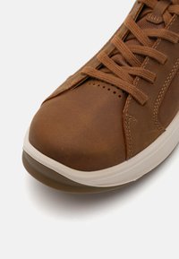 Sneaker din piele maro cu vârf rotunjit, dotat cu șireturi maro și un panou lateral texturat. Talpa este de culoare crem cu nervuri subtile.