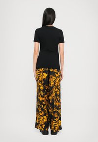 Person with long dark hair wearing a black short-sleeve top and black wide-leg pants with bright yellow floral patterns, standing facing away.