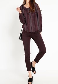 Vínová halenka s potiskem, dlouhými rukávy, kulatým výstřihem a volným střihem, doplněná tmavě vínovými úzkými kalhotami a černými otevřenými klínovými sandály.